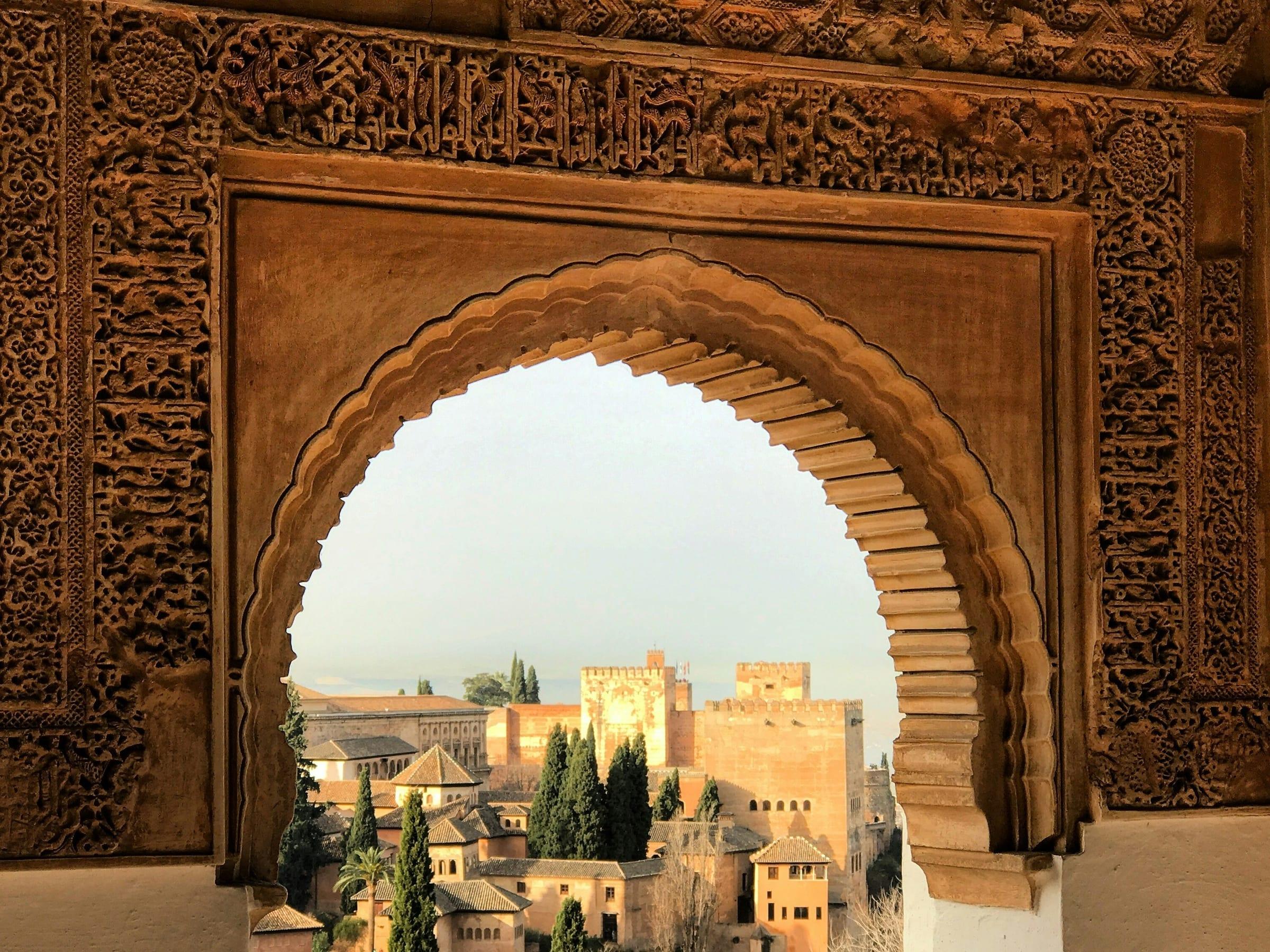 The city of Granada seen through a carved Nasrid horseshoe arch, the Alhambra's own towers rising among cypress trees below.