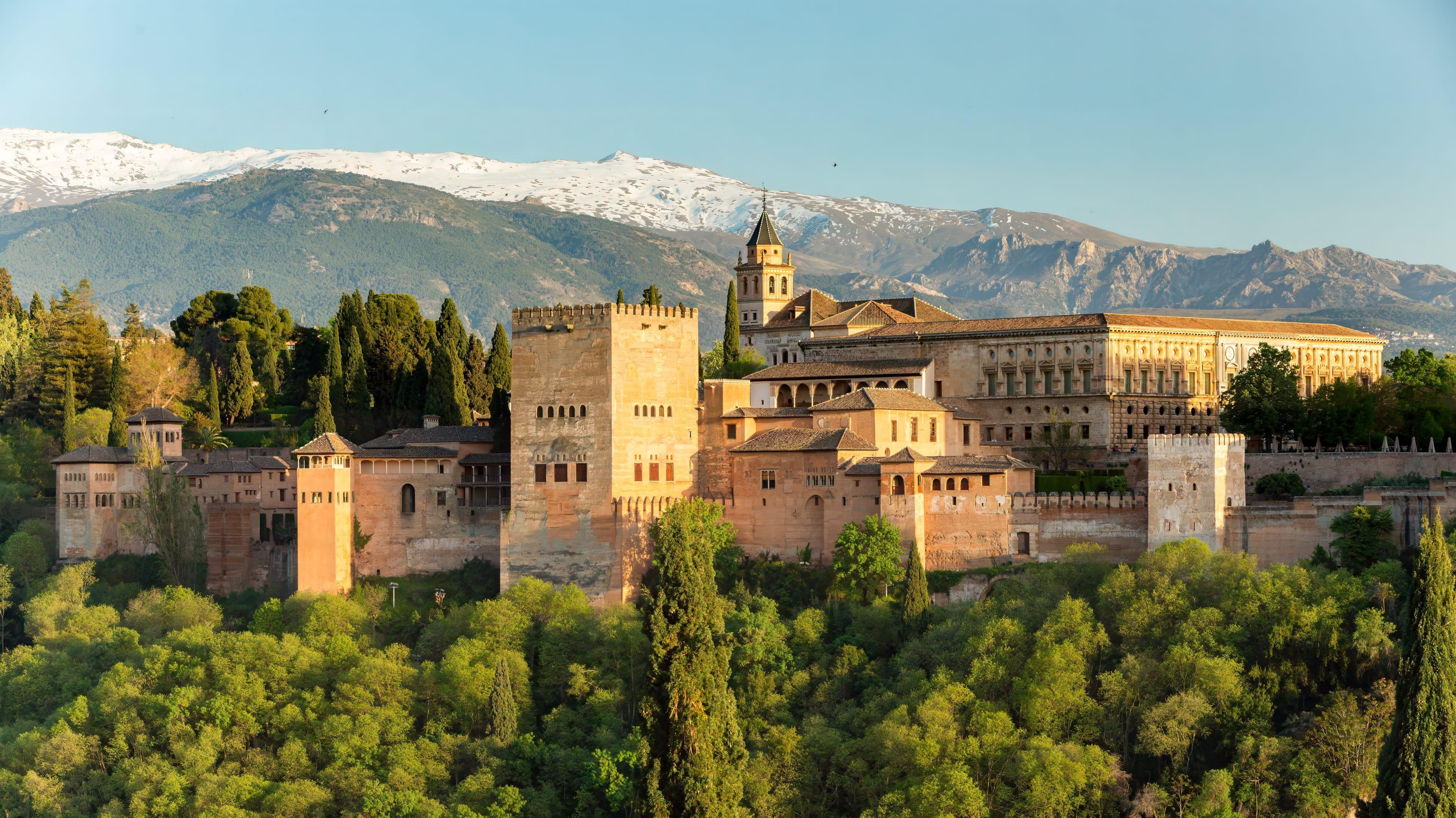 The Alhambra on its wooded hill above Granada, with the snow-covered peaks of the Sierra Nevada rising behind it in late afternoon light.